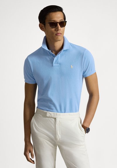 Hellblaues Poloshirt mit strukturierter Oberfläche, mit kurzem Kragen und kleinem Logo. Kombiniert mit cremefarbenen Hosen und einer Armbanduhr.