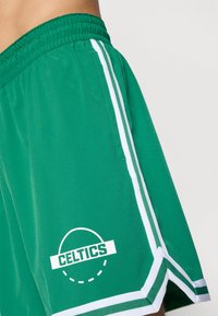 Grønne atletiske shorts med hvide sidestriber og et trykt "CELTICS" logo. Har en snoretræk i taljen og et glat, letvægtsstof.