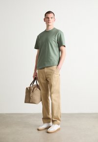 T-shirt vert à manches courtes, pantalon beige et baskets blanches. Porte un sac de sport texturé marron clair avec une fermeture éclair et des poignées.