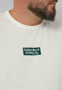 Biała bawełniana koszulka z okrągłym dekoltem, z zielonym prostokątnym logo z napisem "Subskrybuj Naturę" w wyraźnej, bezszeryfowej czcionce.
