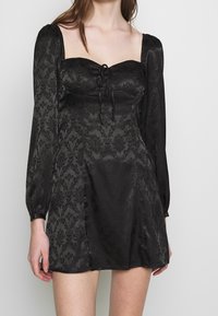 Černé šaty s dlouhými vypouklými rukávy, čtvercovým výstřihem a předním zavazováním. Obsahují jemný damaskový vzor a rozšířený lem. Hladká textura.