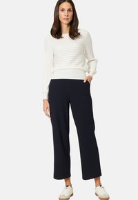 Weißer Ripp-Pullover mit langen Ärmeln, kombiniert mit marineblauen, weit geschnittenen Hosen. Das Model trägt weiße Sneakers mit beigen Akzenten. Einfaches Design, keine Muster.