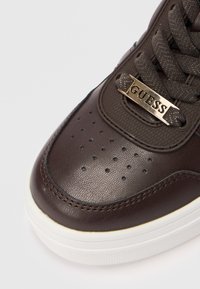 Donkerbruine sneakerneus met perforaties, geprofileerde veters door een gouden metalen plaat met de gravure "GUESS" en een dikke witte zool.