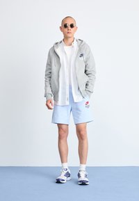 Siv hooded sweatshirt, bela črtasta majica, svetlo modri kratki hlače in sončna očala. Tenisice so v mešanih barvah s strukturiranim dizajnom.