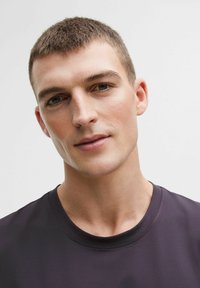 Černé tričko s kulatým výstřihem vyrobené z hladkého a lehkého materiálu. Detailní záběr obličeje osoby se světlou pletí a krátkými vlasy. Neutrální pozadí.