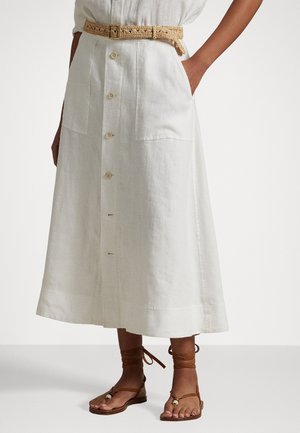 Jupe midi en lin blanc avec des boutons sur le devant, deux poches et une ceinture tissée beige. Des sandales marron avec des lanières autour des chevilles complètent la tenue.