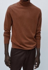 Uomo che indossa un maglione a collo alto marrone aderente e pantaloni marroni scuri su uno sfondo semplice chiaro.