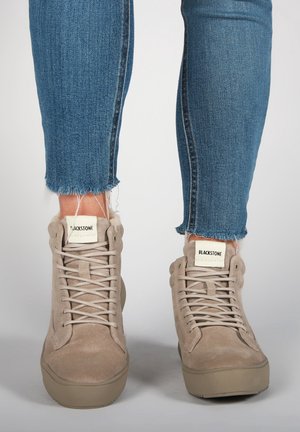 Beżowe zamszowe wysokie sneakersy Blackstone z sznurowadłami, noszone z niebieskimi wąskimi jeansami ze strzępiącym się dołem na jednolitym jasnym tle.