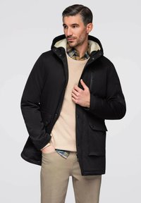 Chaqueta negra aislada con capucha y bolsillos frontales. Se lleva sobre un suéter beige y una camisa a cuadros. Presenta una textura suave y un diseño sutil.