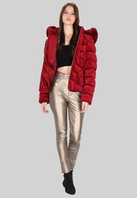 Červená prošívaná bunda s kapucí, s prošívaným vzorem. Komplet doplněný o přiléhavé kalhoty z metalicky zlaté látky a černé ponožky. Jednoduché pozadí.