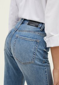 Woman wearing blue denim jeans with back pocket and black Stradivarius label, paired with a white long-sleeve shirt with rolled cuffs.
