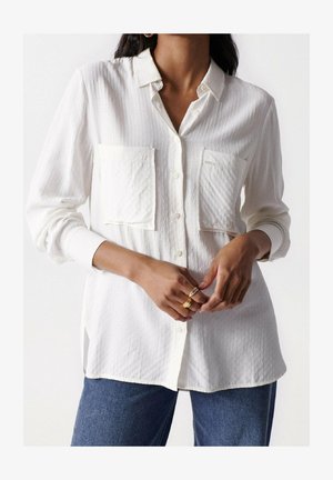 Valkoinen nappikauluspaita, jossa on kaksi rintataskua, teksturoitu kangas ja hieman ylisuurikokoinen istuvuus, yhdistettynä sinisiin farkkuihin.