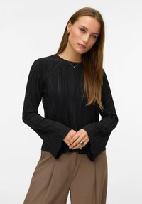 Žena s dugom kosom koja nosi crni plisirani dugi rukav i smeđe hlače, stisnutih ruku ispred sebe na bijeloj pozadini.