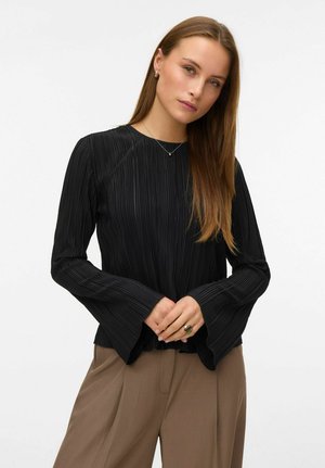 Žena s dugom kosom koja nosi crni plisirani dugi rukav i smeđe hlače, stisnutih ruku ispred sebe na bijeloj pozadini.