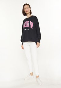 Svart sweatshirt med "BROOKLYN" i rosa bokstäver och "NEW YORK" underneath, tillsammans med vita slim-fit byxor och vita sneakers.