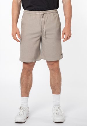 Man draagt beige short met trekkoord, witte sokken en witte sneakers, staat tegen een effen witte achtergrond.