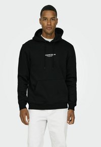 Sort hoodie lavet af en bomuldsblanding, med en kengurulomme, snoretræk i hætten og hvid tekst trykt på brystet. Model iført hvide bukser.
