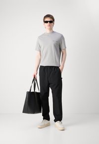 Grå t-skjorte med korte ermer og logo, svarte elastiske bukser, svart totebag og hvite sneakers med beige såler, båret av en person.