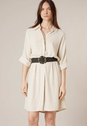 Femme portant une robe-chemise beige mi-longue aux genoux avec des manches retroussées et une large ceinture noire avec une grande boucle ronde.