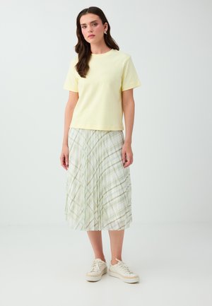 Femme portant un t-shirt jaune pâle à manches courtes, une jupe midi à carreaux blancs et verts, des baskets blanches, debout devant un fond uni clair.