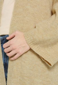 Helle beige gestrickte Strickjacke mit weicher Textur; hat weite Ärmel und eine offene Vorderseite, kombiniert mit einem hellen Oberteil und Jeans.