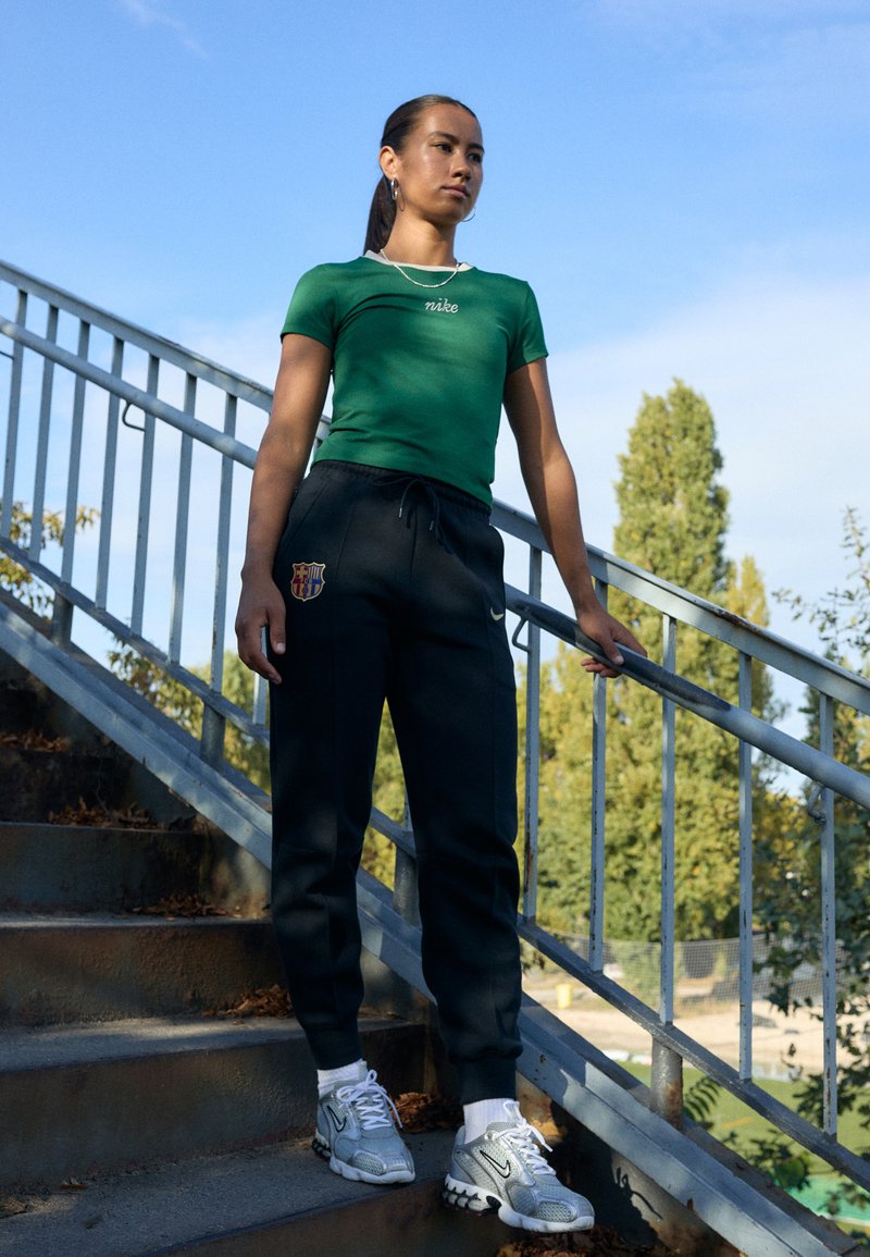 T-shirt verde aderente con logo "nike", abbinato a pantaloni sportivi neri con stemma del Barcellona e scarpe da ginnastica argento.