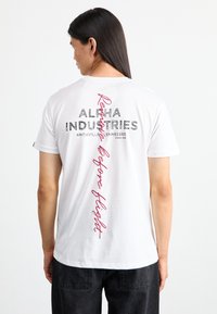 Weißes Baumwoll-T-Shirt mit dem Text "ALPHA INDUSTRIES" in Schwarz und einem roten handschriftlichen Akzent mit der Aufschrift "Reservieren vor dem Flug" auf der Rückseite.