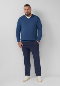 Blauwe V-hals trui van een zachte stof, gecombineerd met navy broek en witte sneakers. Neutrale achtergrond, staande houding, handen in de zakken.