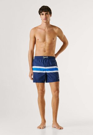 Jonge man staat op blote voeten met één hand achter de rug, draagt donkerblauwe zwembroek met witte en blauwe horizontale strepen.