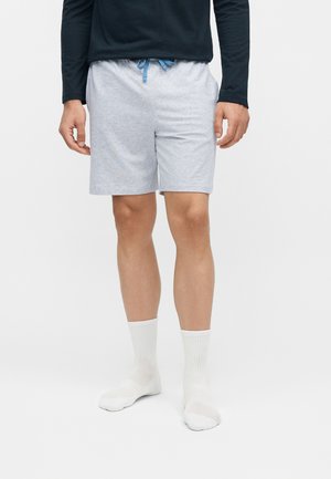 Egy férfi világosszürke húzózsinóros rövidnadrágot, fehér sportzoknit és fekete hosszú ujjú felsőt visel, sík háttér előtt áll.