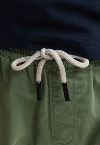 Marineblaues T-Shirt mit glatter Textur, kombiniert mit grünen Shorts. Weiße Kordel mit schwarzen Endstücken, am Bund verknotet.