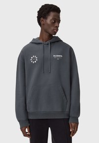 Ciemnoszara bluza z kapturem z miękkiego, teksturowanego materiału. Posiada przednią kieszeń oraz białe logowanie na klatce piersiowej. Kaptur z troczkami. Prosty, casualowy design.