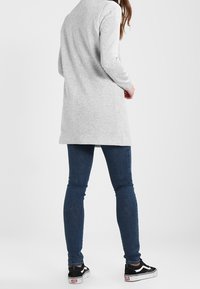 Kobieta ubrana w jasnoszary długi kardigan, ciemnoniebieskie obcisłe dżinsy i czarno-białe trampki, stojąca tyłem na jednolitym tle.