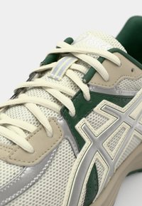 Kraći kadar bež i zelene Asics tenisice s kremastim vezicama, mrežastom tkaninom i srebrnim logotipom na strani.