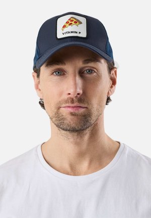 Mann mit leichtem Bart, der eine marineblaue Mütze mit einem Pizza-Stück-Aufnäher trägt, auf dem "Vitamin P" steht, und ein schlichtes weißes T-Shirt, schaut nach vorne.