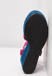 Sandale en daim bleu et rose avec semelle en caoutchouc noire à motifs, placée verticalement contre un mur d’angle blanc et un sol blanc.