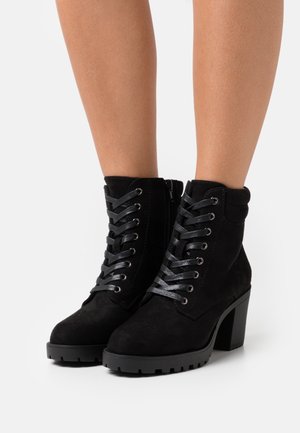 Bottes chevilles en daim noir à lacets avec talons épais portées sur des jambes nues sur fond blanc.