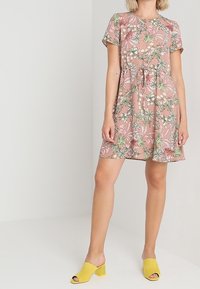 Vestido floral en rosa con patrones verdes y blancos, de mangas cortas y con un lazo en la cintura. Combinado con tacones amarillos destalonados.