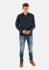Sudadera azul marino con cremallera y un pequeño logo bordado, usada sobre una camiseta azul clara, combinada con jeans azules y botas marrones con cordones.