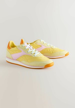 Żółte trampki z różowymi akcentami, pomarańczowym obcasem, białymi sznurówkami i teksturowanymi nakładkami; charakteryzują się klasycznym sportowym designem z gumową podeszwą.