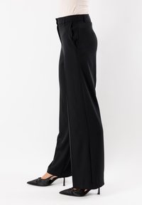 Pantaloni neri su misura lunghi con una vestibilità dritta, caratterizzati da pieghe, una trama liscia e una cintura in vita. Indossati con tacchi neri a punta.