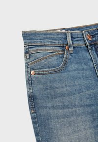 Blå denimjeans med forlomme, møntlomme, bæltestrop og kobbernitter på en hvid baggrund.