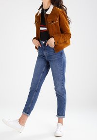 Bruine suède jas met een witte nepbonten kraag, twee borstzakken, gecombineerd met hoog getailleerde blauwe spijkerbroeken en witte sneakers.