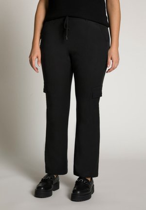 Donna che indossa pantaloni cargo neri con tasche frontali e mocassini chunky neri, in piedi davanti a uno sfondo chiaro semplice.
