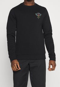 Felpa nera con maniche lunghe, scollo rotondo, polsini e orlo a coste. Presenta un design ricamato colorato sul petto. Tessuto morbido.