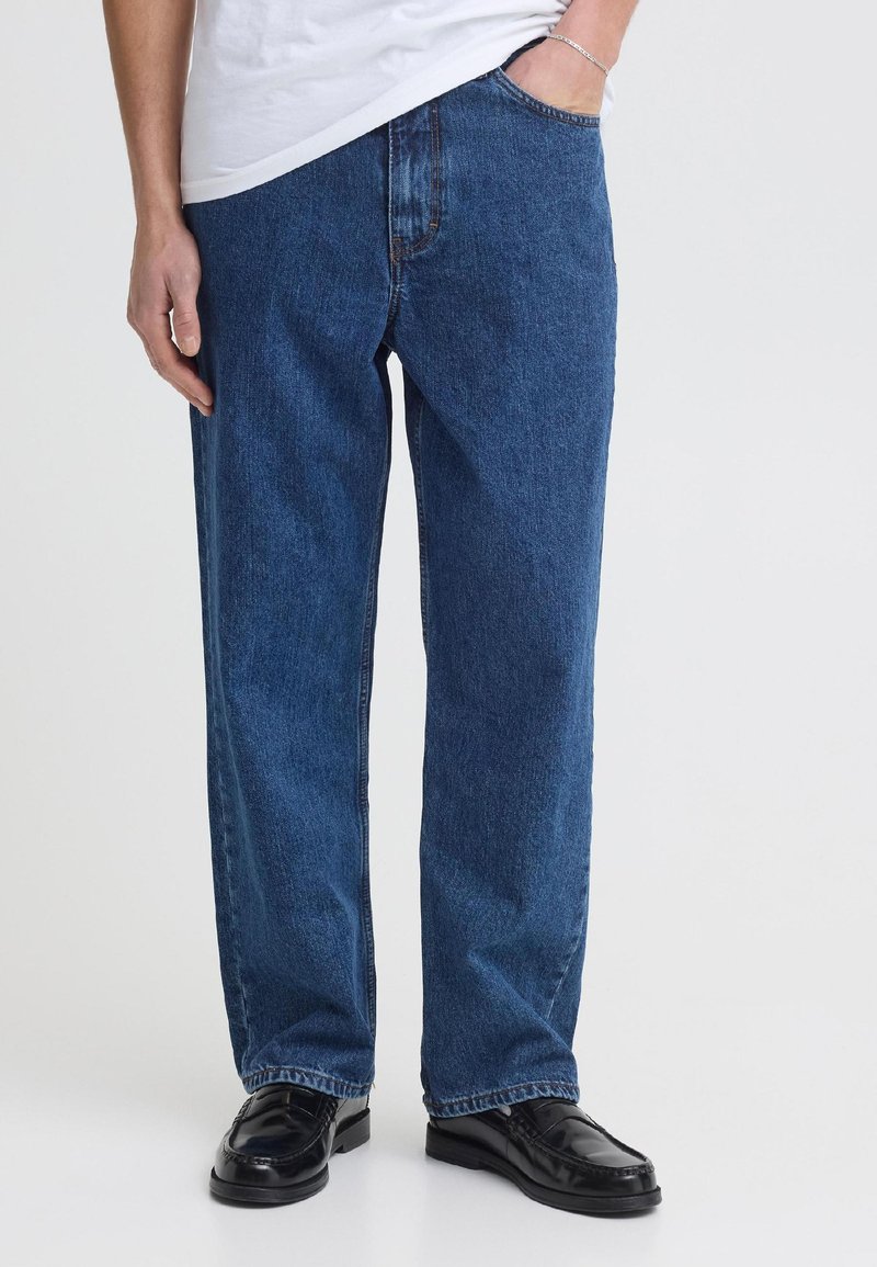 Mann trägt gerade blaue Jeans, schwarze Lederschuhe, weißes T-Shirt, linke Hand in der Tasche und rechte Hand ruht neben dem Bein.