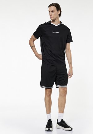 Mężczyzna stojący z ręką na biodrze, ubrany w czarną odzież sportową z napisem „TRUST YOURSELF”, białe skarpetki i czarno-białe buty sportowe na białym tle.