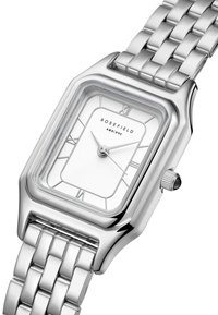 Srebrni pravokutni sat s bijelim brojčanikom, rimskim brojevima i metalnom narukvicom. Ima glatki stakleni poklopac i bočnu krunicu za podešavanje.