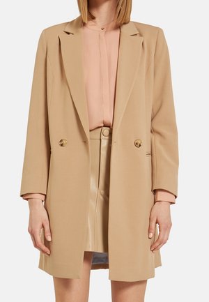 Vrouw draagt een beige dubbelgeknoopte blazer over een lichtroze blouse en een beige lederen rok, staat met de armen ontspannen langs haar zij.