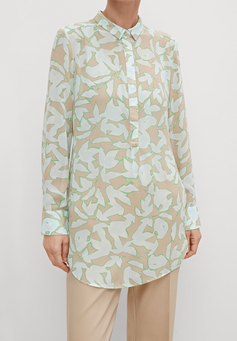 Blusa a maniche lunghe in tessuto leggero e trasparente. Presenta una base beige con motivo floreale in verde pastello e bianco, colletto classico e chiusura con bottoni.